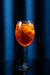 A glass of orange cocktail with ice cubes and orange slices on a dark glossy surface with a blue curtain background