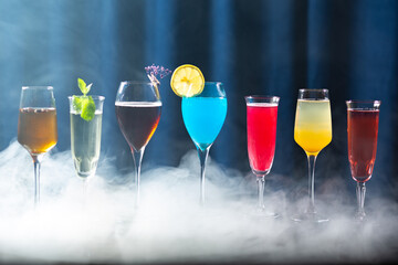 A row of colorful cocktails in elegant glasses with smoke or mist around them, on a reflective surface with a dark blue curtain background