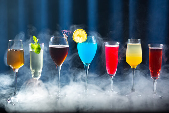 A row of colorful cocktails in elegant glasses with smoke or mist around them, on a reflective surface with a dark blue curtain background