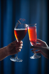 Two hands holding glasses with red cocktails in a celebratory toast against a deep blue background. One glass is garnished with a small bunch of dried flowers and a cinnamon stick, adding a festive to