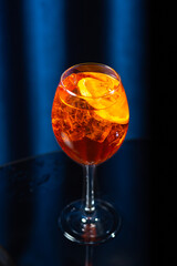 A glass of orange cocktail with ice cubes and orange slices on a dark glossy surface with a blue...