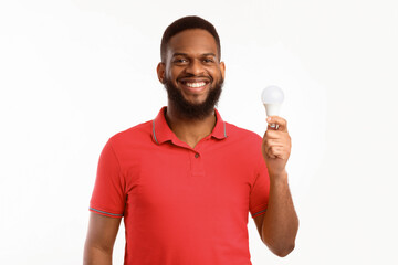 Happy Black Guy Showing Energy-Saving Lightbulb Standing Over White Studio Background, Posing...