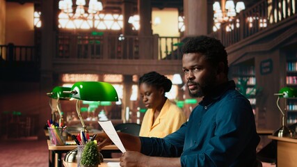 Determined pupil preparing for exams at the campus library, using educational resources like books, notes and research papers for complete information. Quiet environment for productivity. Camera A.
