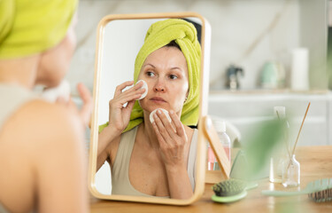 Focused and calm woman with towel wrapped around head attentively removing makeup with cotton pads...
