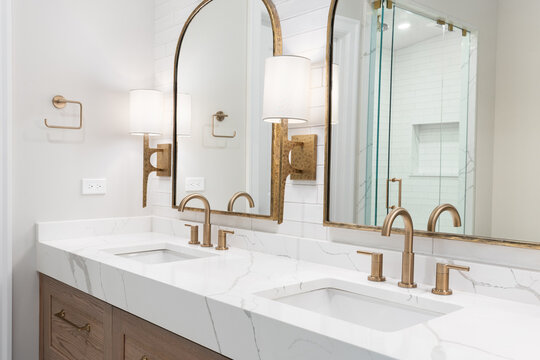 A bathroom detail with gold faucets, light fixtures, and mirrors, a wood cabinet, large marble slab countertop, and a subway tile shower. - Powered by Adobe