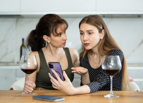Upset ladies in their middle ages gazing at the phone screen regretfully while drinking a glass of wine