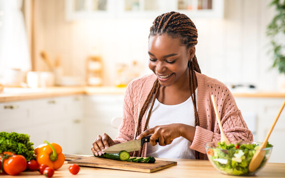 Happy young black woman cooking fresh vegetable salad in kitchen, chopping cucumber. Smiling millennial african american lady preparing lunch at home, enjoying eating healthy food, copy space