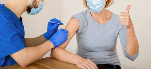Mature woman in face mask approving of covid-19 vaccination, showing thumb up gesture during...