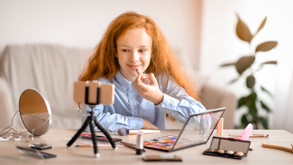 Beauty Blog For Teenagers. Young beautiful blogger doing makeup, recording cosmetics product review, applying lipstick and looking at camera. Girl filming vlog for her channel with cellphone on tripod