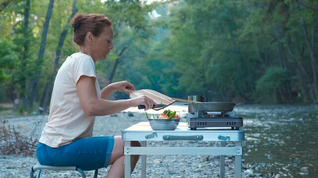 A woman cooks at a campsite by the river. Woman preparing fresh vegetables at riverside portable table, surrounded by lush greenery