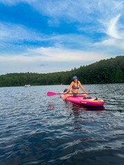 A woman sits on a bright pink paddleboard in the middle of a calm lake, smiling brightly while...