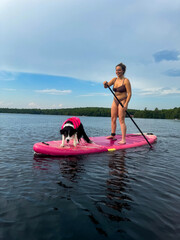 A woman stands and paddles on a pink paddleboard, with her dog standing beside her in a life jacket. They are on a calm lake having a great time.