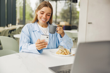 A young woman is happily enjoying her delicious breakfast while checking her phone in a cozy cafe setting