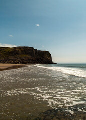 A scenic view of a beach with waves crashing on the shore, a cliff in the background, and a clear blue sky.