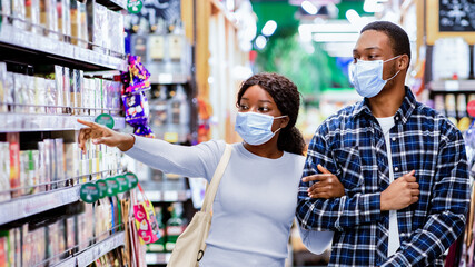Shopping for groceries during covid lockdown. Young black couple in face masks walking through supermarket rows with cart, pointing at shelves, choosing food products together