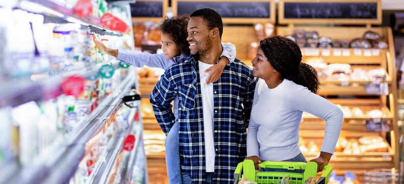 Portrait of happy black family with trolley shopping together at grocery store. Millennial African American parents with lovely daughter picking food, choosing milk products at big mall