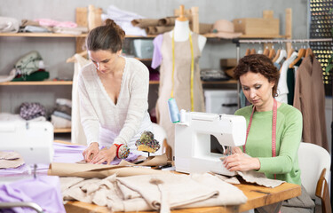 Two skilled female tailors focusing on creating custom garments in small workshop, cutting fabric...