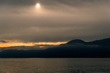 Lake Garda is captured in late afternoon light with a cloudy sky. The sun appears as a soft glowing...