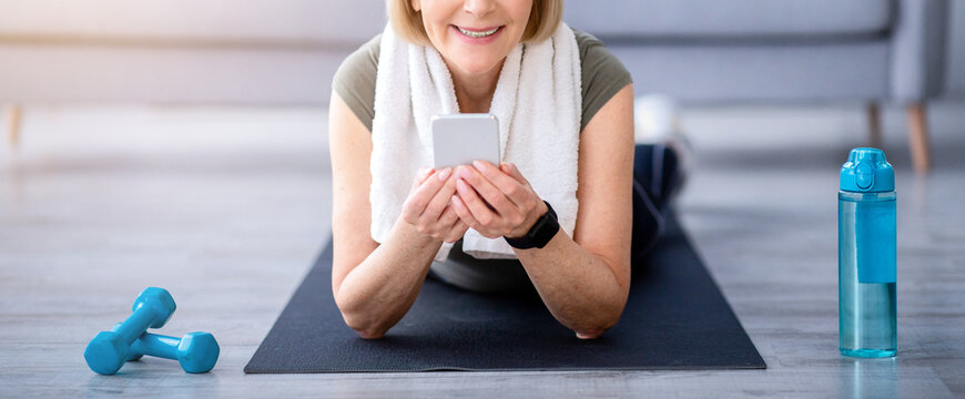 Cheerful senior woman resting on yoga mat, browsing internet on smartphone at home. Happy mature lady taking break after home fitness or yoga, watching online sports video on mobile device