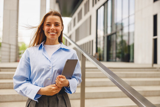 A woman in a blue shirt holds a tablet outside a modern office building, exuding confidence and professionalism