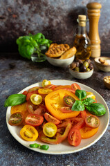 Front view of colorful sliced tomatoes with fresh basil, olive oil bottle and wooden pepper mill on dark blue brown background