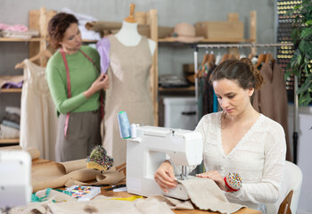 Working in sewing workshop - female tailor sewing on sewing machine, another tailor trying on dress on a mannequin