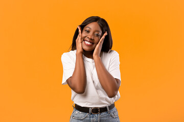 Emotional black lady in white t-shirt smiling and touching her face over orange studio background,...
