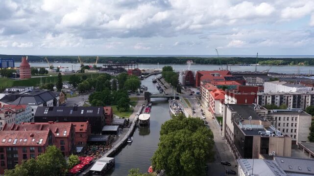Panoramic aerial view of Klaipėda, Lithuania, showing the river port, red-roofed historic buildings, pedestrian waterfront, boats, and the Baltic Sea in the distance under a partly cloudy sky.