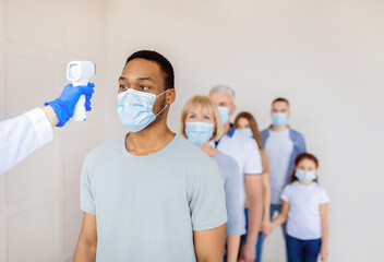 Coronavirus immunization medical campaign concept. Black man in face mask standing in line at clinic, doctor measuring his temperature with infrared thermometer before covid-19 vaccination, copy space