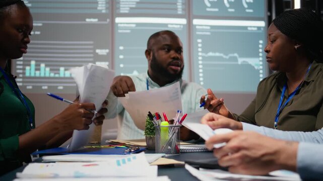 Close up of diverse specialists evaluating data numbers on archived files, exchanging reports and insights to establish clear enterprise objectives in strategy room late at night. Camera A.