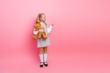 Charming schoolgirl holding teddy bear on pink background while gesturing happily with playful...