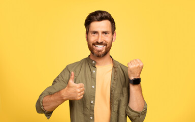 A middle aged man in his 40s shows his smartwatch with a thumbs up gesture. He has a beard and is wearing casual clothing, smiling brightly against a vibrant yellow background.