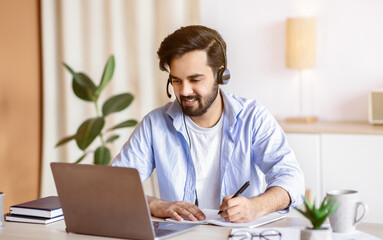 Fototapeta premium Online Education. Young Arab Guy In Headset Watching Webinar On Laptop Computer And Taking Notes, Eastern Millennial Man Enjoying Distance Learning, Sitting At Desk At Home Office, Free Space