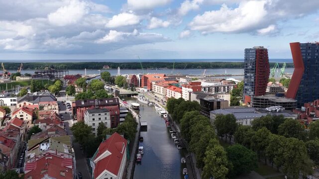 Panoramic aerial view of Klaipėda, Lithuania, showing the river port, red-roofed historic buildings, pedestrian waterfront, boats, and the Baltic Sea in the distance under a partly cloudy sky.