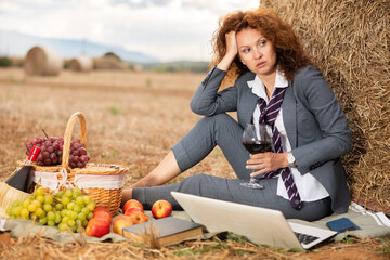 Tired business woman in stylish gray suit with white shirt and loosened tie sitting on blanket with picnic basket and laptop, leaning against bale of straw on mown field, relaxing with wine in retreat