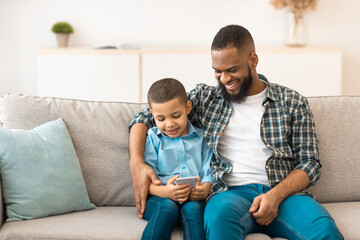 Black Boy And His Dad Using Mobile Phone Playing Games Online Sitting Together On Couch And Browsing Internet In Living Room Indoor. New Application For Smartphone, Family And Gadgets Concept