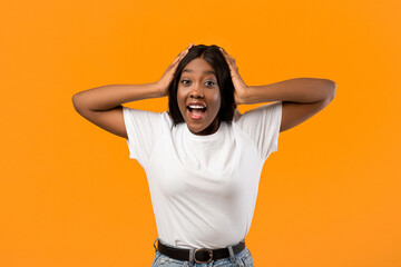 Shocked black lady in white t-shirt raising hands up and touching head over orange studio...