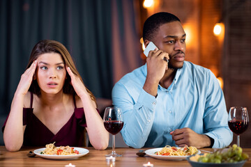 Woman looking unhappy on date while her boyfriend talking on cellphone in restaurant, paying no attention to her and busy using his mobile phone. Multiracial couple dining together at Valentine's day