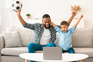 Joyful Black Father And Son Watching Soccer Game Online On Laptop Computer Cheering Favorite Team Sitting On Couch At Home. Football Fans Concept. Selective Focus