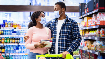 Millennial black couple in protective masks checking their weekly food shopping list, buying...