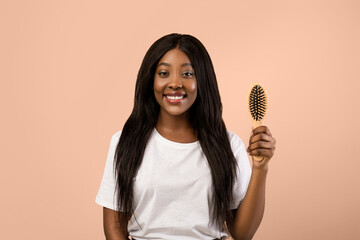 Attractive young black woman holding wooden hair brush and smiling over beige studio background. Happy african american lady with beautiful long hair enjoying her new wooden hair brush