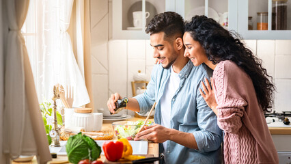 Modern muslim couple cooking healthy food in kitchen together, middle eastern spouses preparing vegetable meal for lunch at home, husband adding seasoning oil to salad in bowl, free space