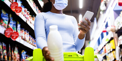 Young black woman in disposable face mask holding smartphone, going through shopping list at...