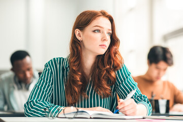 Fototapeta premium Learning Process Concept. Portrait of Pensive Ginger Lady Sitting At Desk Taking Notes Writing in Exercise Copybook And Looking Aside At Window, Thinking. Young Person Learning New Stuff In Highschool
