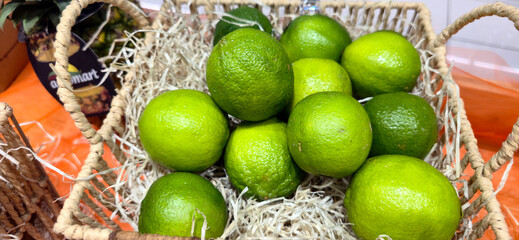 Fresh green limes arranged in a wicker basket filled with straw, displayed for sale on an orange...