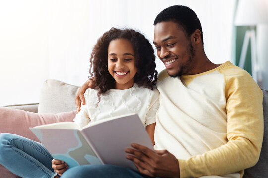 Family lifestyle, leisure activity concept. Happy black father and daughter reading book together at home, copy space. Beautiful african american family dad and child reading fairy tales
