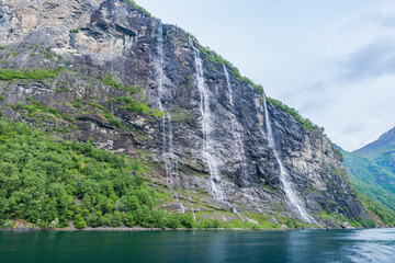 Angled view of the massive Seven Sisters waterfall and surrounding mountains in the UNESCO landmark, lush green tree-lined fjord named Geirangerfjord, in the Nordic village, Geiranger, Norway at day