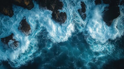 Aerial view of powerful ocean waves crashing against dark jagged rocks creating white foam and turbulent blue water