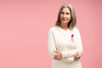 Obraz premium Happy, smiling cheerful senior woman wearing white sweater with pink ribbon posing with crossed arms