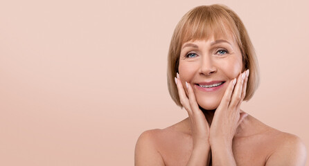 Lovely senior lady with perfect skin touching her face and smiling at camera over blue studio...